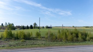 Mavi gökyüzünün altında yeşil tarlaları, ağaçları ve elektrik hatları olan kırsal alan..