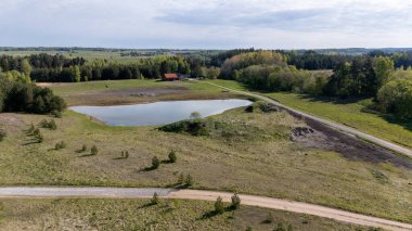 Bulutlu bir gökyüzünün altında tarlalar ve ormanlarla çevrili küçük bir göleti olan kırsal bir arazinin havadan görünüşü..