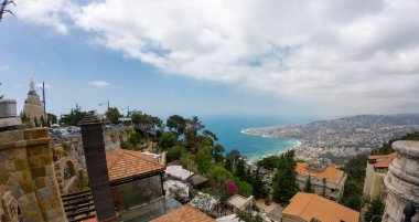 Açık mavi gökyüzü, yemyeşil yemyeşil ve denize bakan binaları olan panoramik bir kıyı şehri manzarası. Lübnan