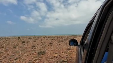 Bulutlu bir gökyüzünün altında okyanusun yakınındaki kayalık bir çöl yolunda giden bir araba..