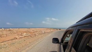 Bir araba, açık bir gökyüzünün altındaki çöl manzarası boyunca kırsal bir yolda gidiyor..