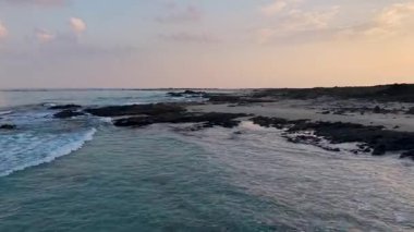 Gün batımında turkuaz okyanus, yumuşak dalgalar, kumlu sahil, ve pastel gökyüzü, sakin kıyı manzarası. Sokotra, Yemen.