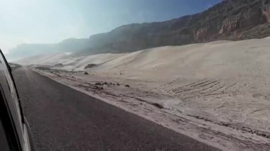 Soluk kum tepeleri ve engebeli kayalık dağların yanında asfalt karayolu manzaralı çöl manzarası, bir araçtan bakıldığında lastik izleri ve rüzgarın süpürdüğü dokular görülüyor. Sokotra, Yemen.