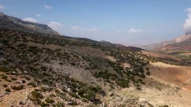Mavi gökyüzünün altında dağınık yeşil çalıları olan kurak kayalık tepelerin geniş panoramik manzarası engebeli dağları ve uzak bir vadiyi gün ışığında gösteriyor. Sokotra Adası.