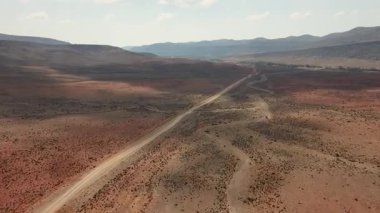 Kızıl-kahverengi tepeler ve sisli bir gökyüzünün altındaki uzak dağlarla uçsuz bucaksız bir çöl vadisini kesen uzun toprak yolun havadan görünüşü. Sokotra, Yemen.