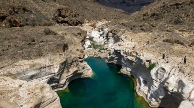 Kurak bir dağ manzarasında dar bir turkuaz nehir havuzunu çerçeveleyen katmanlı kireçtaşı kayalıkları olan Rocky Canyon 'da birkaç yürüyüşçü gün ışığı altında kenarda duruyor. Kalysan Kanyonu, Sokotra.