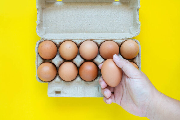 Man's hand is picking up a brown egg in a carton box. On a yellow background.