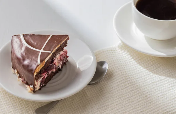  Bir parça lezzetli çikolatalı kek, beyaz tabakta çilekli ve bir fincan kahveli. Tahta masanın üzerine güneş ışığı. Kahvaltı konsepti.