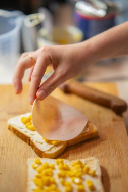 Hızlı bir yemek hazırlayan bir elin yakın çekimi, tereyağlı tostun üzerine ince bir dilim jambon ve tahtadan yapılmış mısırla..