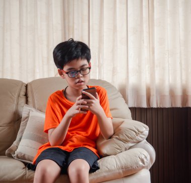 Gülümseyen Asyalı çocuk koltukta otururken cep telefonuyla çizgi film izliyor. Çocuk evde dinlenirken daktilo ve cep telefonu kullanıyor..