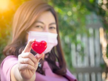 Grip virüsünden korunmak için tıbbi koruyucu yüz maskesi takan kadın Covid-19 Coronavirüs salgını sırasında kırmızı kalbi karantina için tutuyor.. 