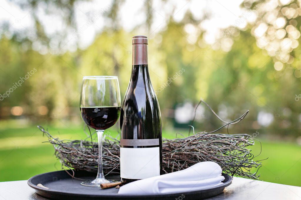 A bottle of red wine with a full glass stands on the grass in the evening light