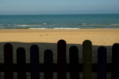 Ahşap çit, altın bir kumsal ve sakin bir turkuaz deniz manzarası sağlıyor. Yaz kaçışını ve deniz tatillerini çağrıştırıyor.
