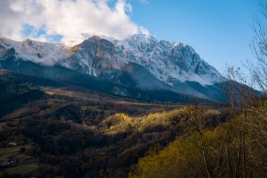 Mavi gökyüzünün altında karla kaplı dağ sıraları, aşağıdaki vadide altın sonbahar ağaçlarının parlak bir kısmını aydınlatan güneş ışığı.