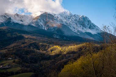 Berrak bir gökyüzünün altındaki karlı dağ zirvelerini vadideki altın sonbahar ormanını aydınlatan güneş ışınlarını gösteren dramatik kış manzarası.