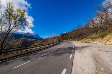 Etrafı ağaçlarla çevrili, verimli doğal bir arazide, sarmal asfalt yol, kırsal bir ahşap çit, ve uzakta görkemli bir kar tepesi.