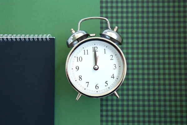 Open spiral notepad with black pages and a beautiful vintage silver alarm clock on a green checkered background. List of goals for the new year