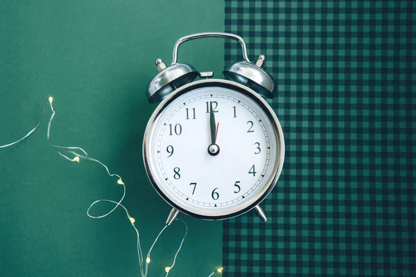 Beautiful vintage silver alarm clock and a glowing garland on a green checkered background. Time concept. Holiday routine