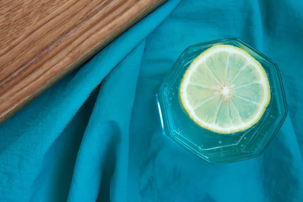 Glass of water with a slice of lemon on turquoise fabric and wooden cutting board. Tropical fruits. Fresh citrus. Top view