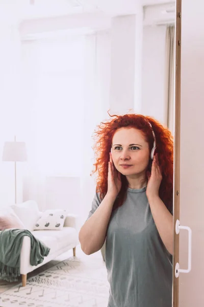 Φωτεινή σγουρή γυναίκα με ακουστικά ακούει μουσική και κοιτάζει την αντανάκλασή της στον καθρέφτη — Φωτογραφία Αρχείου