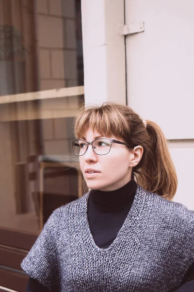 Νεαρή όμορφη γυναίκα με γυαλιά, σε ένα άνετο γκρι-μπλε γιλέκο περνά χρόνο σε εξωτερικούς χώρους — Φωτογραφία Αρχείου