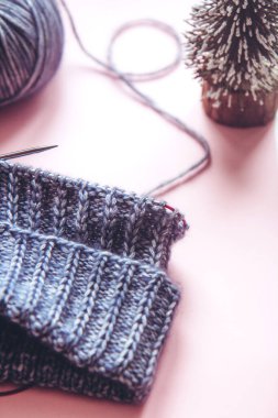 Örgü projesi başladı. Mavi iplik yumağı, örgü iğneleri ve açık pembe arka planda Noel ağacıyla örgü örmek. Yünlü giysiler ören bir kadın.