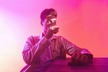 Bir bardak viskiyle turuncu-mor arka planda bir adam.