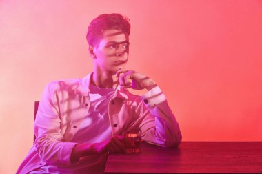 Bir bardak viskiyle turuncu-mor arka planda bir adam.