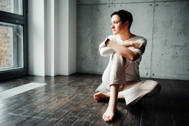 Genç bir kadın modern bir dairede yerde oturuyor ve pencereden dışarı bakıyor.