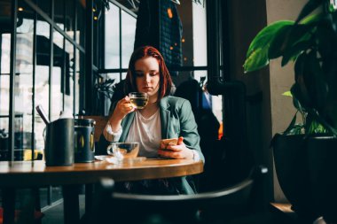 Genç bir kız kafede yeşil çay içiyor ve telefonda bir mesaj okuyor.