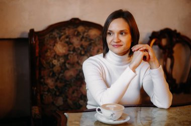 Tarz sahibi bir kadın. Ellerine yaslanıyor ve yan tarafa bakıp gülümsüyor. Bir kafede masada oturuyor..