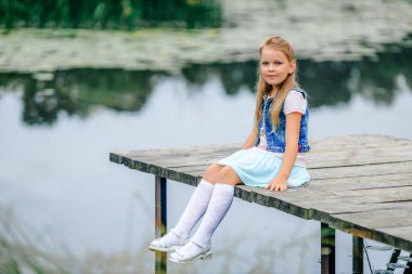 Okul çağındaki güzel bir kız, bir gölün kıyısında ya da bir nehrin kıyısında, bacakları sarkık bir şekilde oturur ve gülümseyerek kameraya bakar.