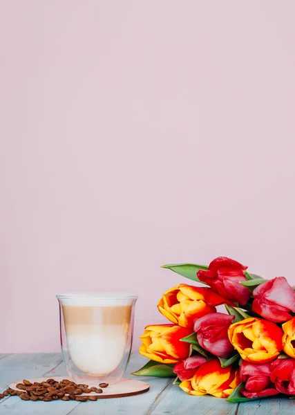 İki duvarlı bir bardakta kapuçino ve bir buket lale ile kavrulmuş kahve çekirdekleri. Fotokopi alanı olan kadınsı pembe arka plan. Yüksek çözünürlüklü görüntü, sığ alan derinliği.