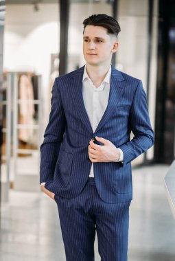 Mavi takım elbiseli ve beyaz gömlekli genç bir iş adamı elini cebinde, diğerini de ceketinin düğmesinde görüyor..