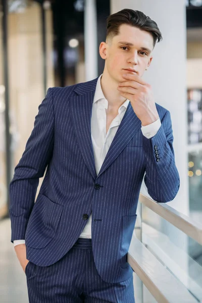 portrait of sexy young businessman in navy blue suit holding chin, looking at camera and posing, holding hand in pockets.