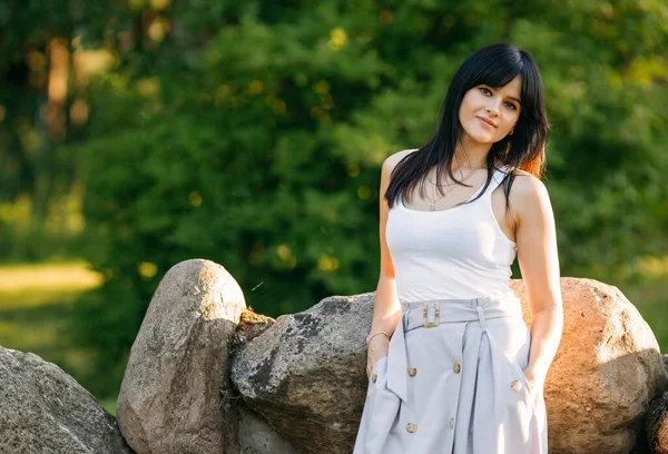 A beautiful brunette girl stands against the background of stones and green trees in the park in the rays of the setting sun