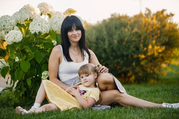 Mother and little daughter are sitting on the grass in the summer park. The girl laid her head on her mothers lap. They look into the camera. Outdoor recreation concept