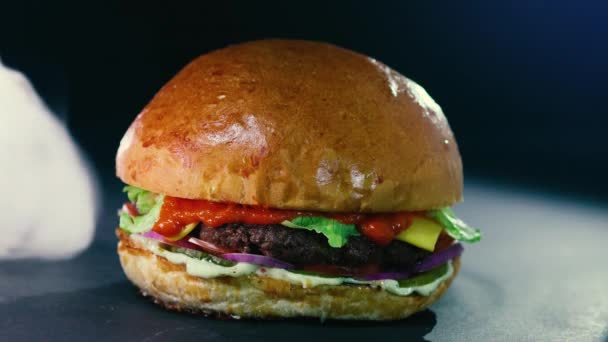 Big mouth-watering burger with meat cutlet, onions, vegetables, melted cheese, lettuce and mayonnaise sauce. Isolated hamburger on a dark background of smoke, close-up view — Stock Video