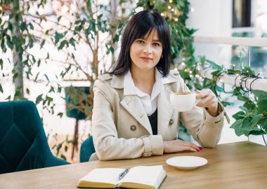 Elinde bir fincan kahveyle bir kafede oturmuş kameraya bakan tatlı bir esmer kız..