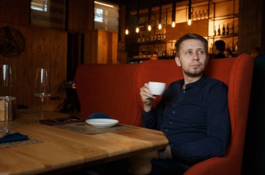 Genç bir adam pahalı bir restoranda elinde beyaz bir fincan kahveyle oturur ve yan tarafa bakar..