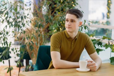 Genç yakışıklı serbest çalışan bir kafede oturuyor, taze kahve içiyor ve başka tarafa bakıyor..