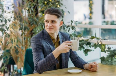 Zarif resmi kıyafetler içindeki mutlu genç bir adam bir otel kafesinde oturur ve öğleden sonra kahvesini içer. İş sırasında kahve molası..