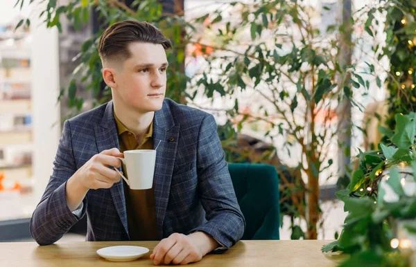 Resmi takım elbiseli konsantre genç bir adam elinde bir fincan kahveyle masada oturur, yan tarafa bakar ve modern bir kafede iş hakkında düşünür..