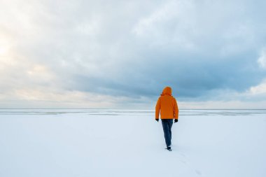 Turuncu ceketli yalnız bir adam karla kaplı bir göle doğru yürür..