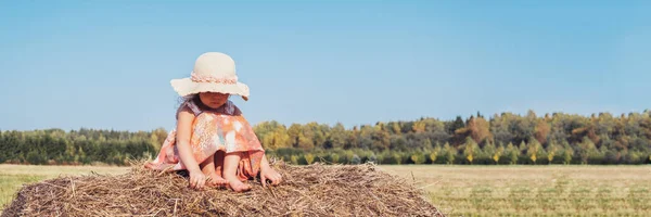 Cute little girl in a straw hat and a summer dress sad, in a village on a big hay stack on the field. Summer and warm, country rest. Banner.