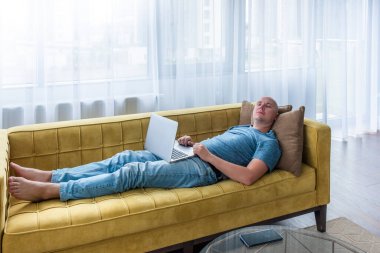 Dizüstü bilgisayar kullanırken mavi tişörtlü bir adam sarı kanepede uyuyor. Uzaktan çalışma, evden çalışma.