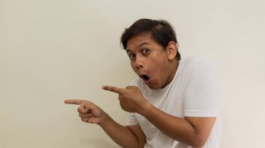 Beyaz tişörtlü Asyalı genç Malezyalı adamın, izole edilmiş beyaz arka planda boş bir yeri gösteren yüz ifadesi ve şaşkınlığı..