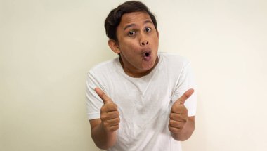 Beyaz tişörtlü Asyalı genç Malezyalı adamın, izole edilmiş beyaz arka planda boş alana çift parmak işareti göstermesi komik bir yüz ifadesi. Seçici odak.