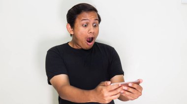 Siyah tişörtlü Asyalı genç Malay 'ın beyaz arka planlı akıllı telefonuna bakışları şok edici bir yüz ifadesi. İyi haber konsepti. Seçici odak.