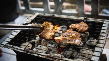 Piknik partisinde sıcak dumanlı kömür ızgarasında pişirilmiş metal maşayla sosla marine edilmiş çiğ kuzu eti tutan eli yakından izleyin. Önplanda seçmeli odak.